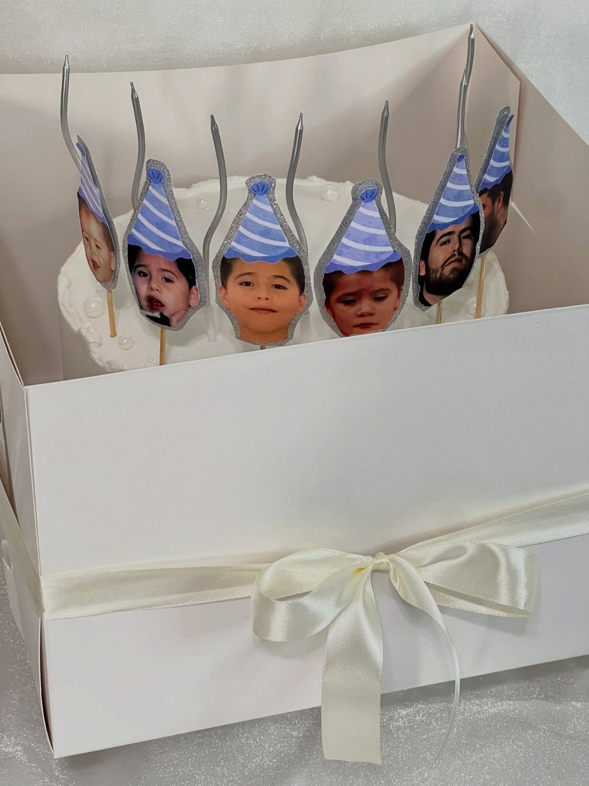 White cake with edible toppers of a child's face wearing blue party hats.