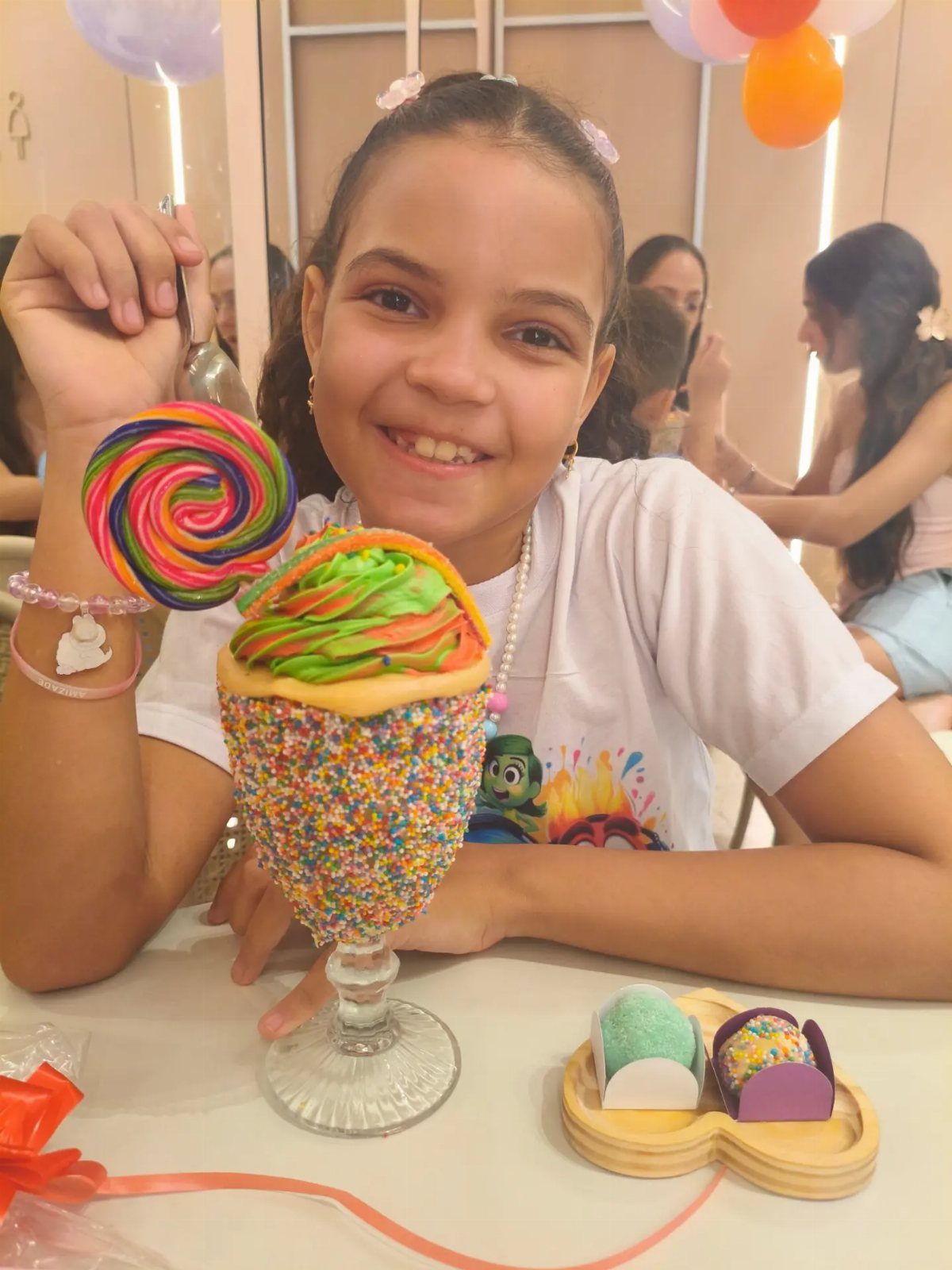 Smiling girl with a colorful rainbow lollipop and sprinkle-covered milkshake dessert.