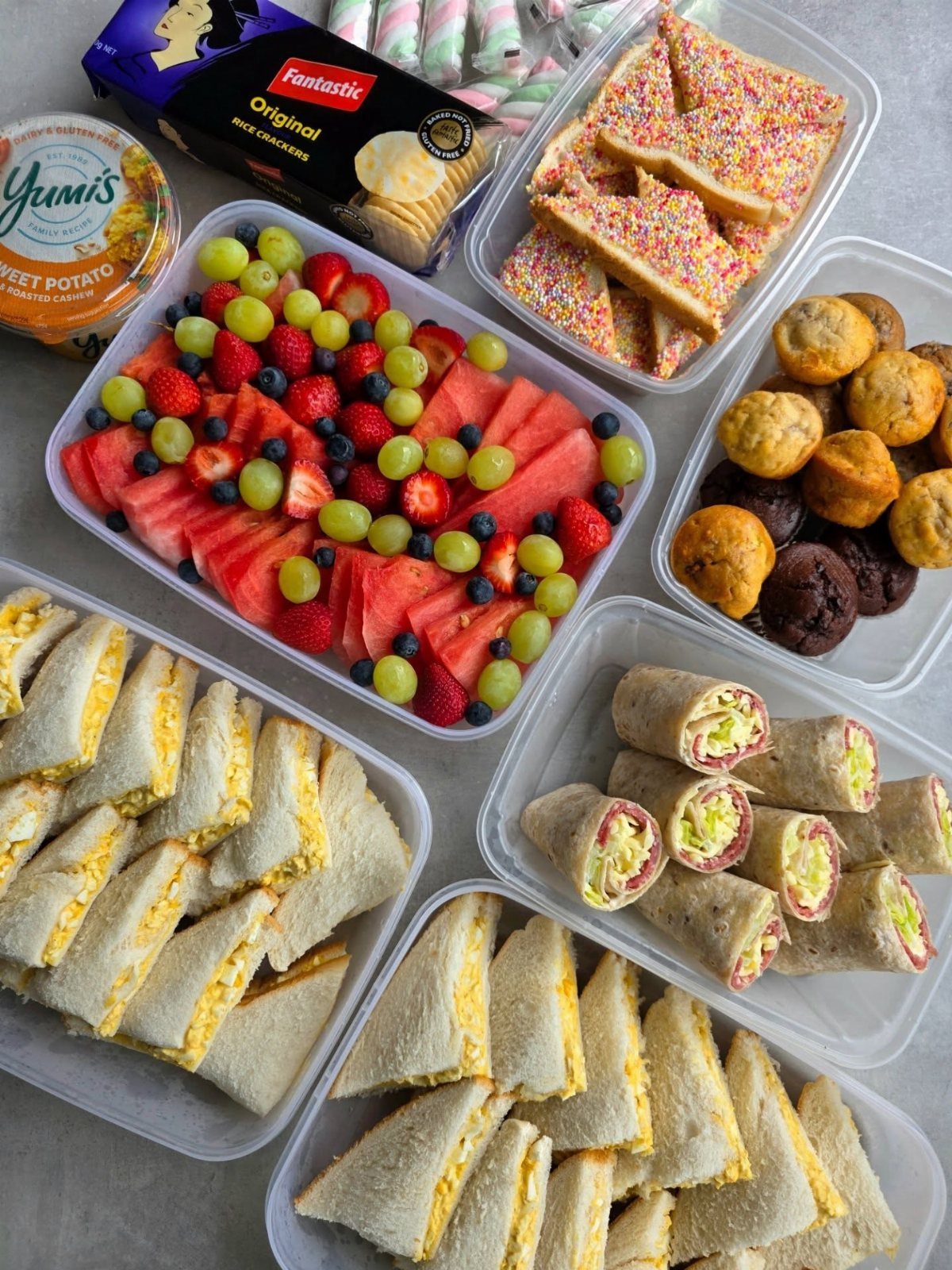 Assorted picnic foods including sandwiches, wraps, fruit, and muffins in plastic containers.