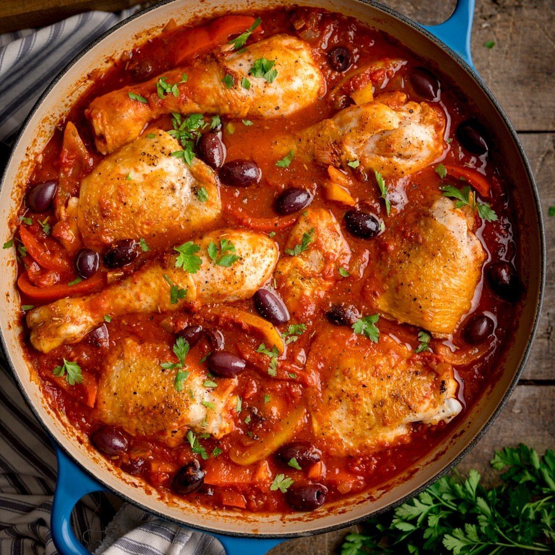 Chicken drumsticks and thighs in tomato sauce with olives and peppers in blue pot.