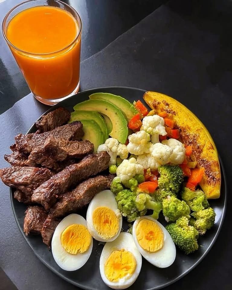 Grilled steak, boiled eggs, avocado, and steamed vegetables on a black plate with juice.
