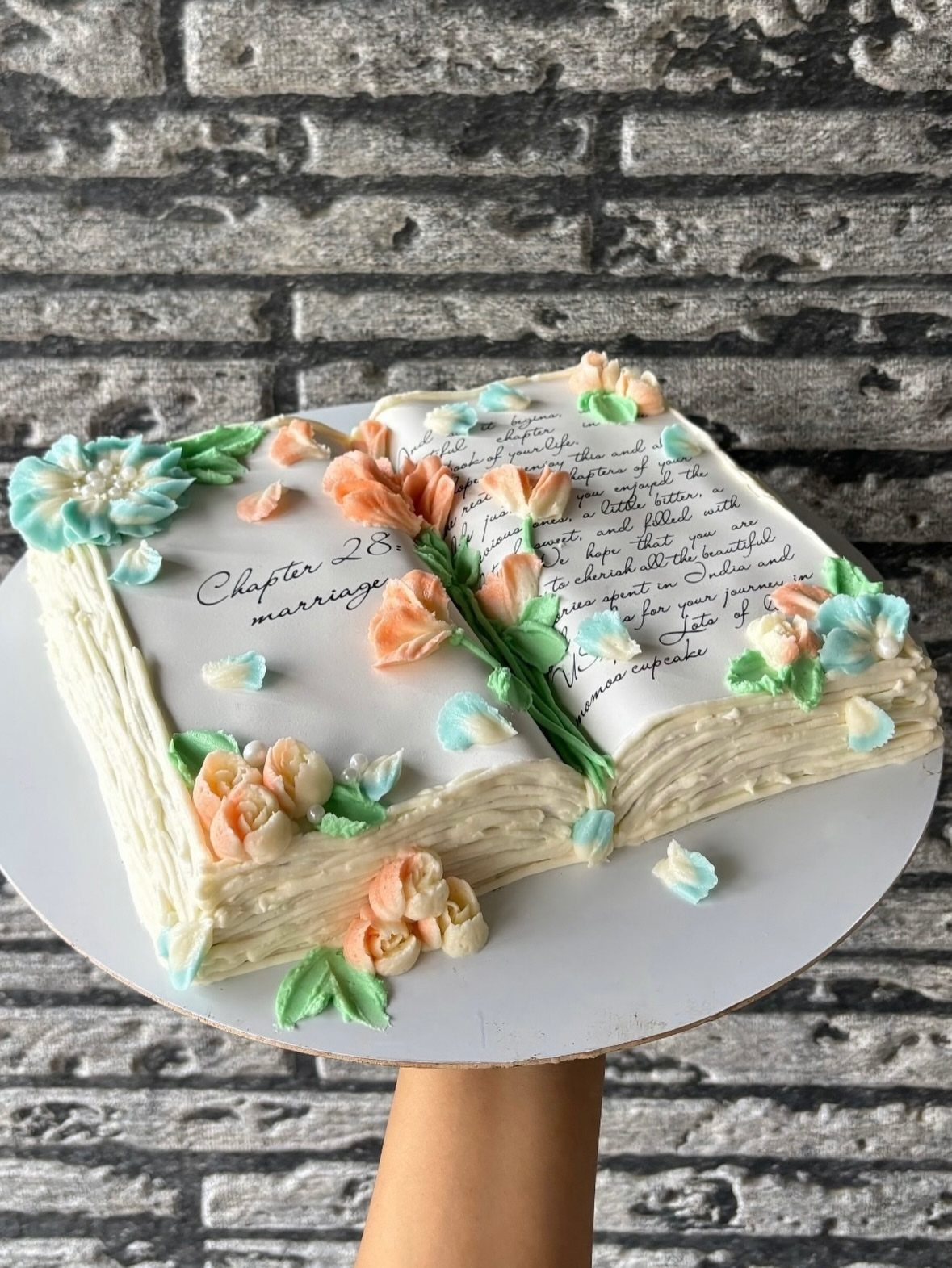 Open book cake decorated with orange and blue buttercream flowers and cursive text.