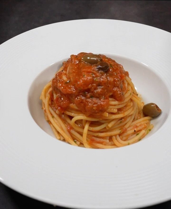 Spaghetti with chunky tomato sauce and green olives in a white bowl.