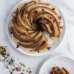 Spiral bundt cake with beige glaze, rose petals, and pistachios on white plate.