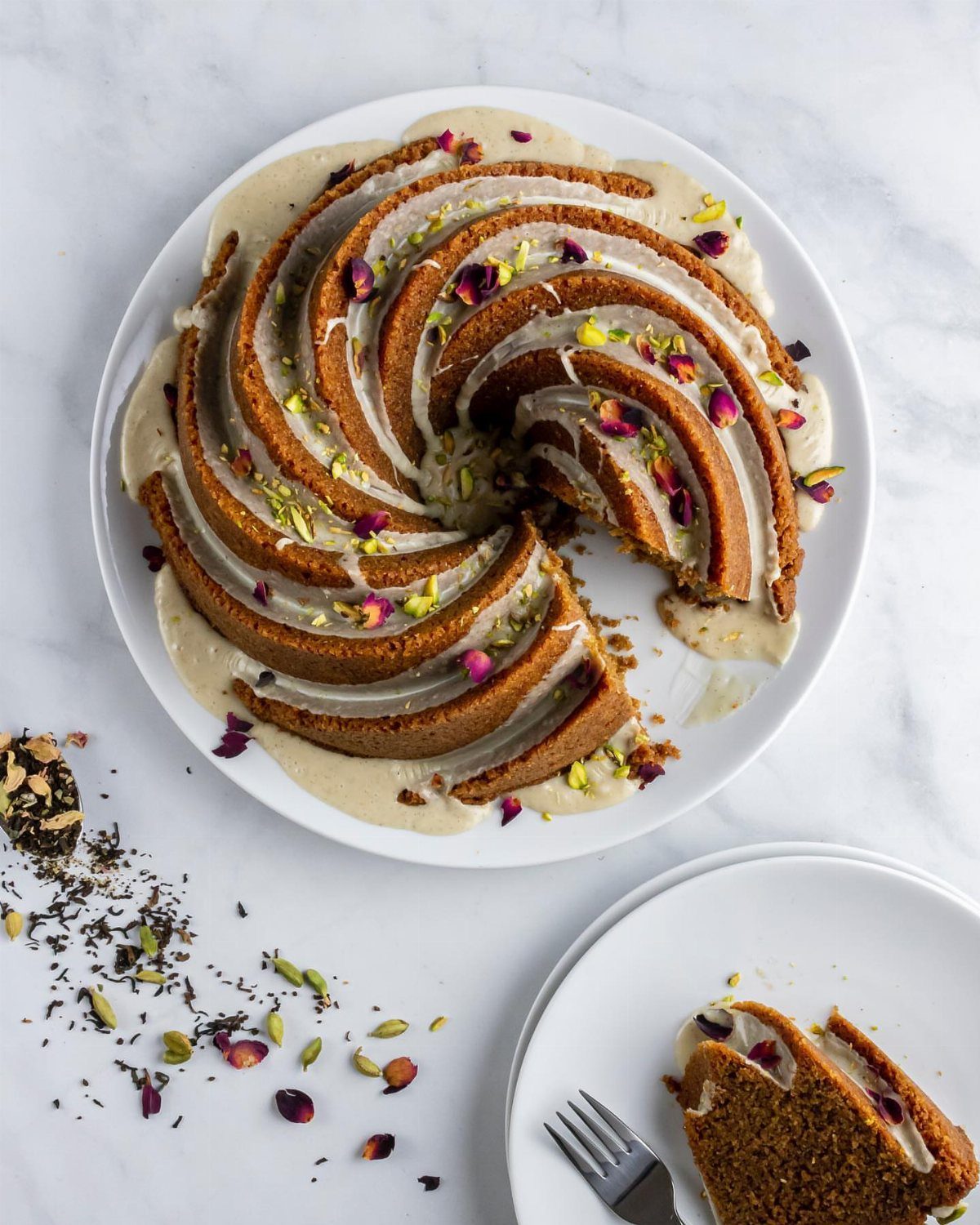 Spiral bundt cake with beige glaze, rose petals, and pistachios on white plate.
